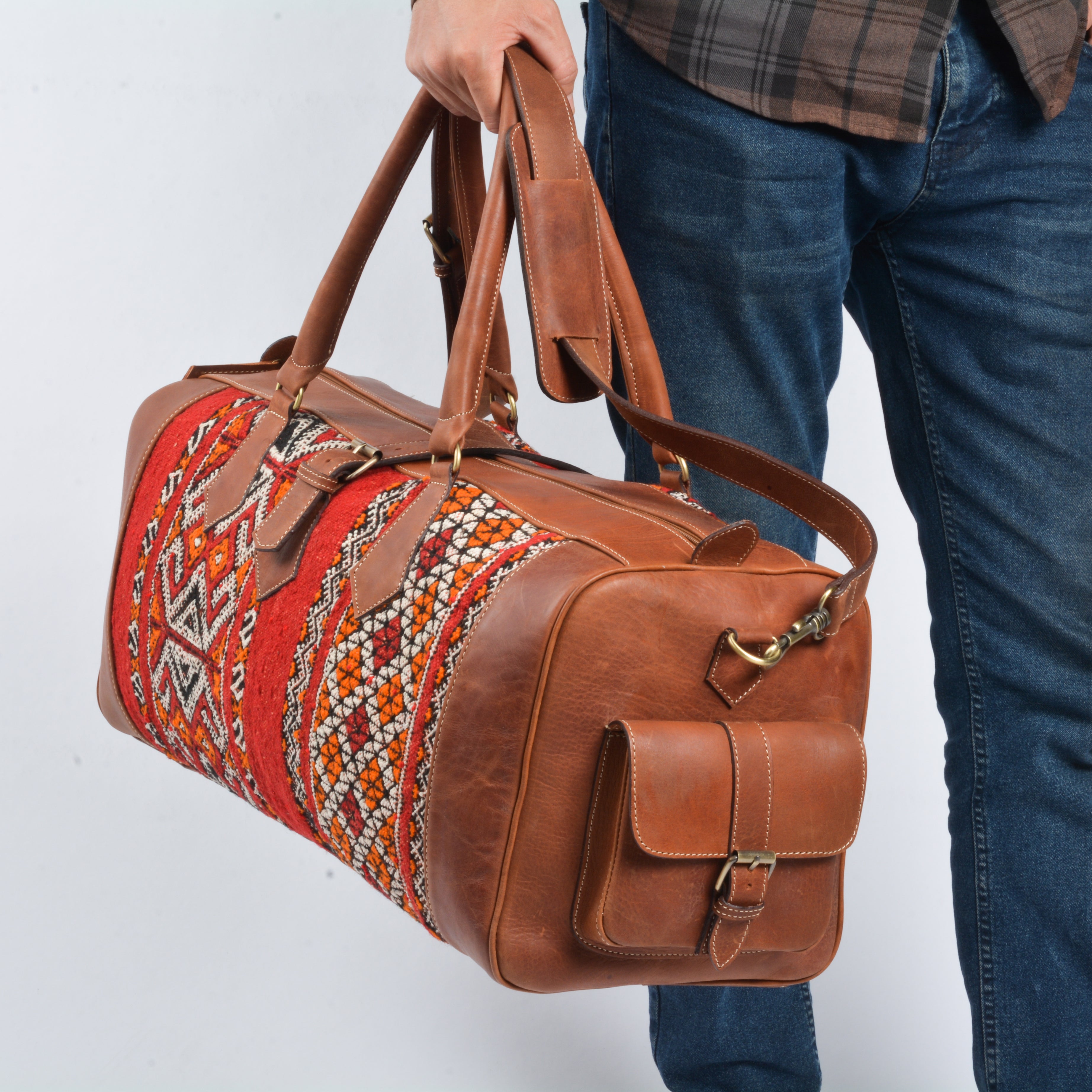 A person carrying a brown leather travel bag with a colorful kilim pattern on the interior lining. The bag has a shoulder strap and handles, and appears to be made of high-grade cotton.