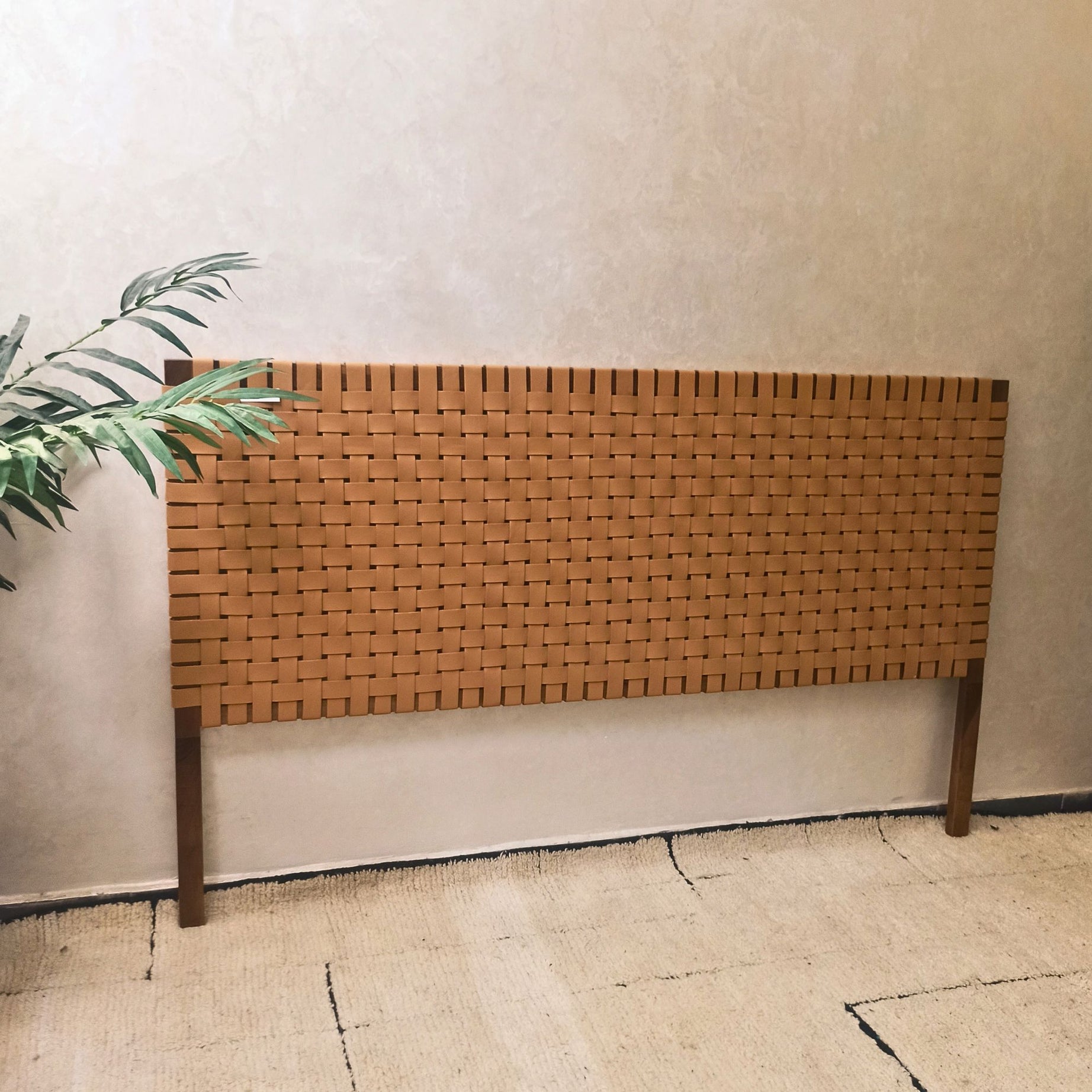 Woven walnut wooden headboard against a plain wall with a plant to the left.